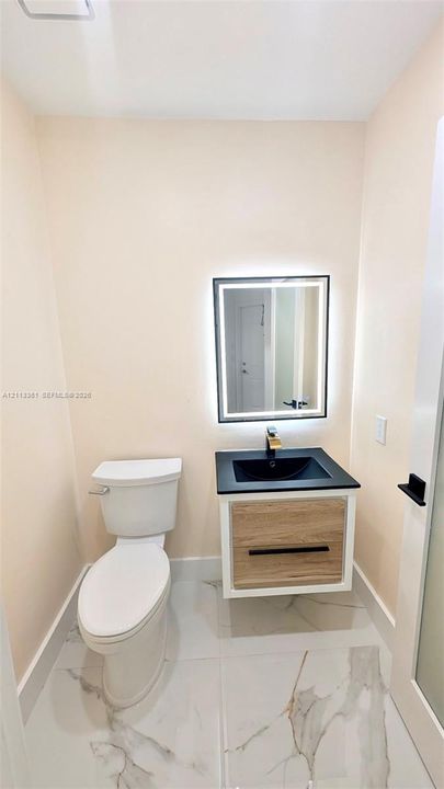 Floating vanity with wood-finish drawer, matte black hardware, stone-surface countertop, integrated vessel sink, and rectangular LED-backlit mirror, complemented by large-format tile flooring