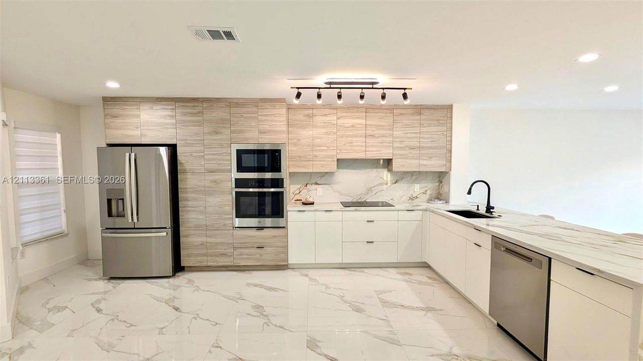 Kitchen featuring two-tone cabinetry, stone-finish countertops, full-height backsplash, stainless steel built-in appliances, and large-format tile flooring