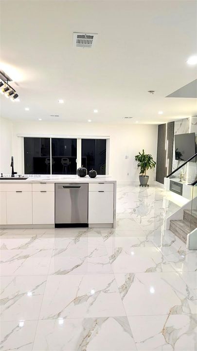 Large-format stone flooring featuring a veined pattern, white flat-panel cabinetry, stainless steel dishwasher, glass-panel staircase railing, and built-in electric fireplace with vertical accent lighting