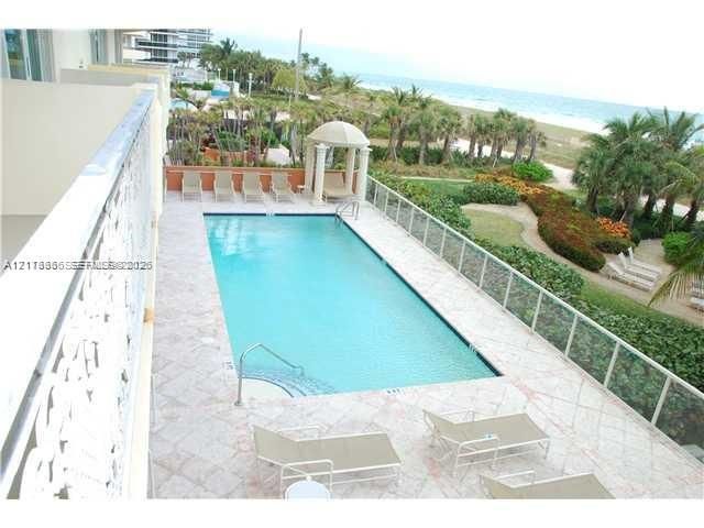 Rectangular swimming pool featuring a tile deck, glass panel perimeter railing, and a columned cabana structure, situated against a sandy coastline backdrop