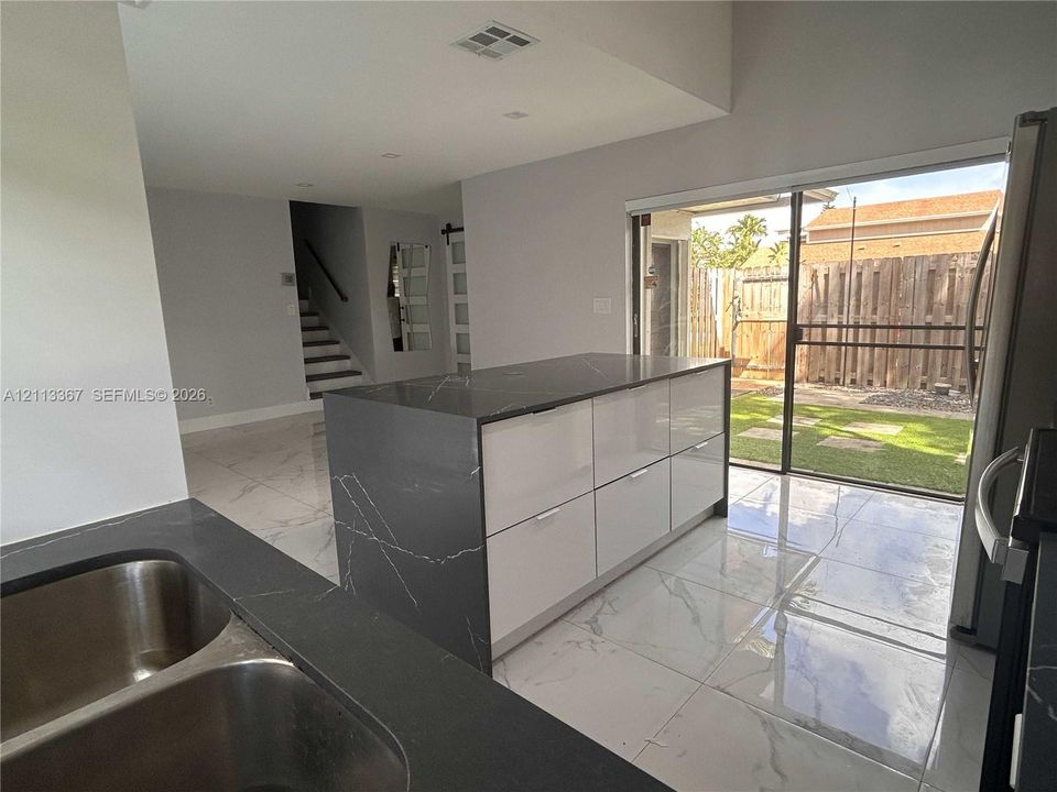 Kitchen featuring a central island with high-gloss cabinetry and a dark stone-finish countertop, dual-basin stainless steel sink, large-format tile flooring, and a sliding glass door