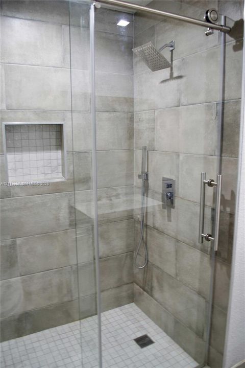 Walk-in shower featuring large-format neutral tile walls, a sliding glass door with stainless steel hardware, a rainfall showerhead, a handheld sprayer, and a built-in recessed niche