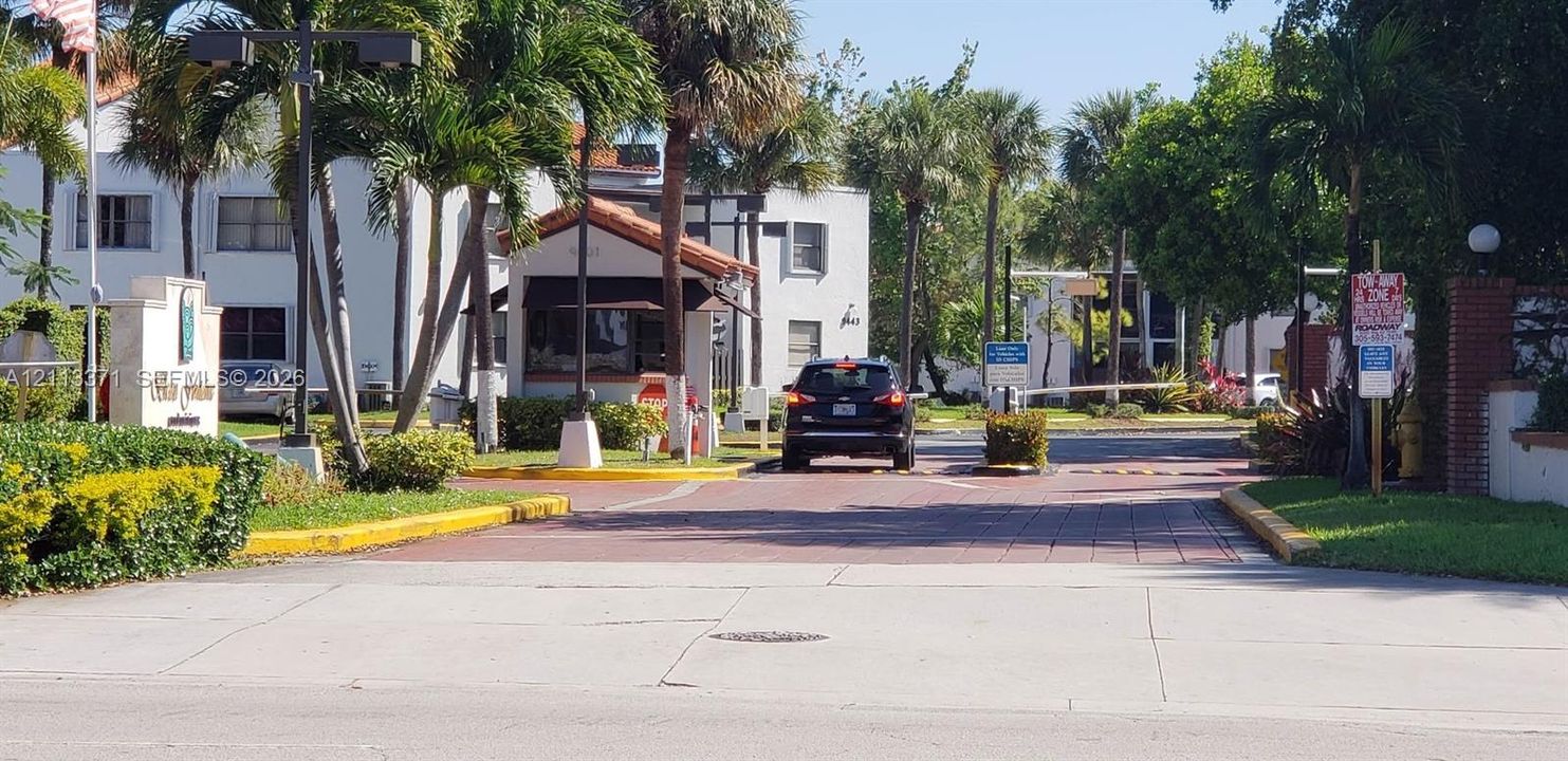 Gated entrance featuring a brick-paved driveway, guard house, mature palm trees, and landscaped perimeter