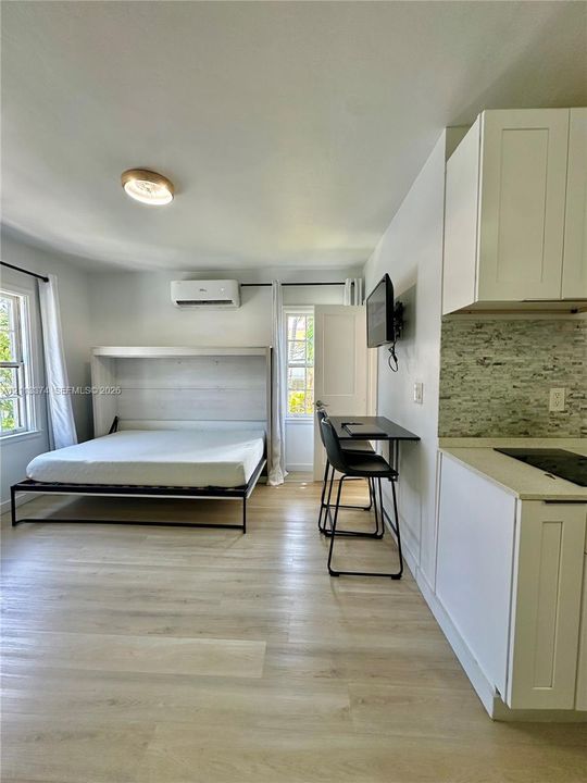 Wood-finish flooring, white shaker-style cabinetry, stone-textured tile backsplash, wall-mounted ductless climate control unit, and built-in wall bed