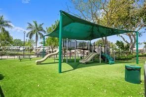 Outdoor recreational area featuring a playground structure with dual slides, protective shade canopy, synthetic turf surfacing, and surrounding mature trees