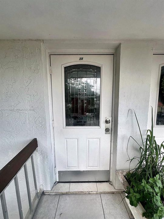 White entry door featuring an arched decorative glass insert with metal caming, a brushed metal kickplate, and a dual-lock system, set within a textured stucco exterior wall