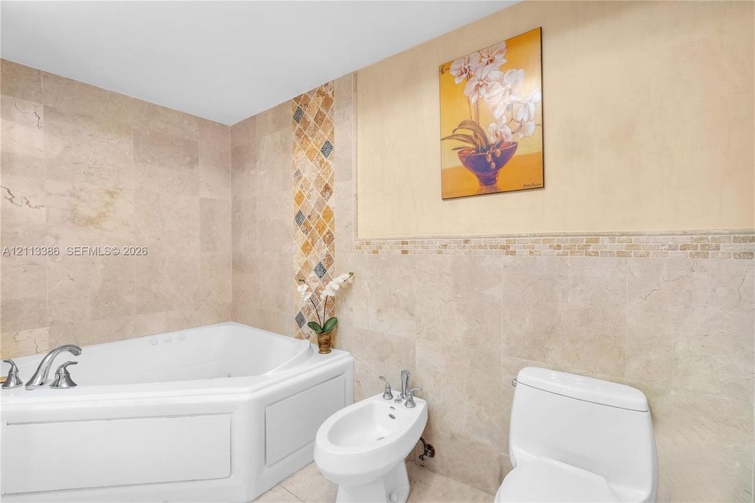 Corner soaking tub with deck-mounted chrome faucet, bidet, two-piece toilet, and floor-to-ceiling stone tile walls with decorative glass and stone mosaic accents