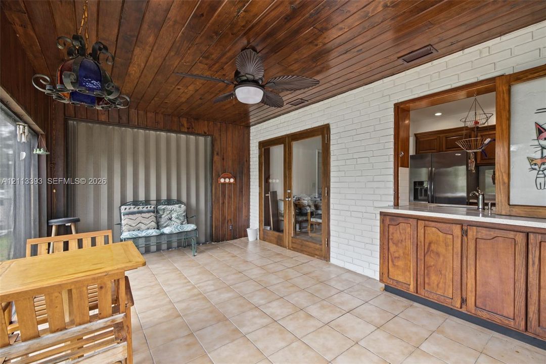 Enclosed patio featuring wood-paneled ceiling, white-painted brick walls, tile flooring, built-in cabinetry with stone countertop, and glass-paneled double doors