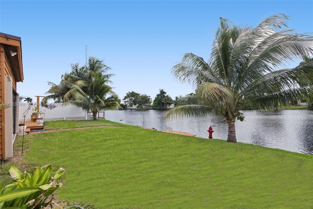 Waterfront lot featuring a grass lawn, established palm trees, wooden decking, and a privacy fence