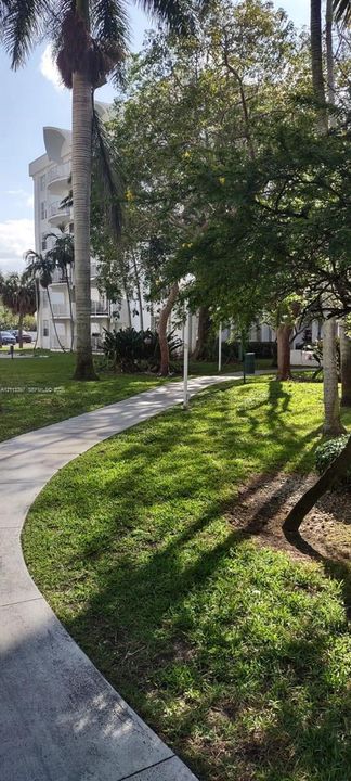 Curved concrete walkway through landscaped grounds featuring mature trees, manicured lawns, and palm trees