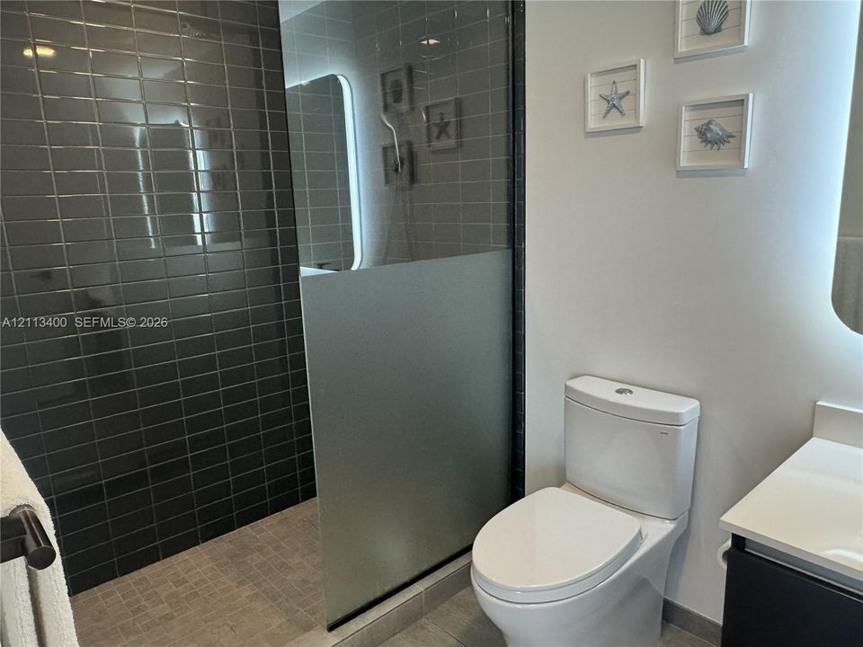 Walk-in shower featuring dark rectangular wall tile, mosaic floor tile, and a partial privacy glass screen, paired with a white porcelain toilet and a vanity with a solid-surface countertop