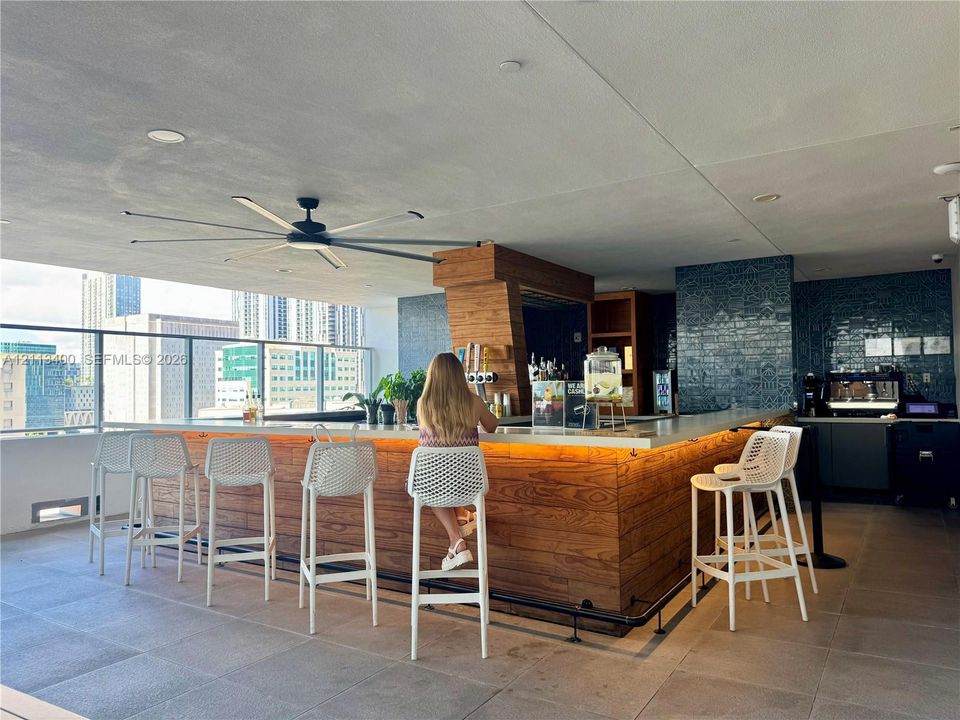 Outdoor bar featuring wood-paneled cabinetry, stone countertop with integrated under-cabinet lighting, glass-tiled backsplash, and a large overhead ceiling fan on a covered terrace