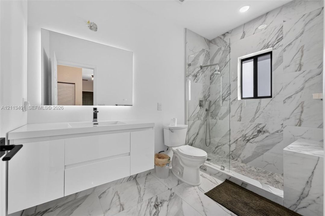 Floating white vanity with integrated sink, expansive LED-backlit mirror, floor-to-ceiling stone-look wall tiles, walk-in glass-enclosed shower, and matching stone-look tile flooring