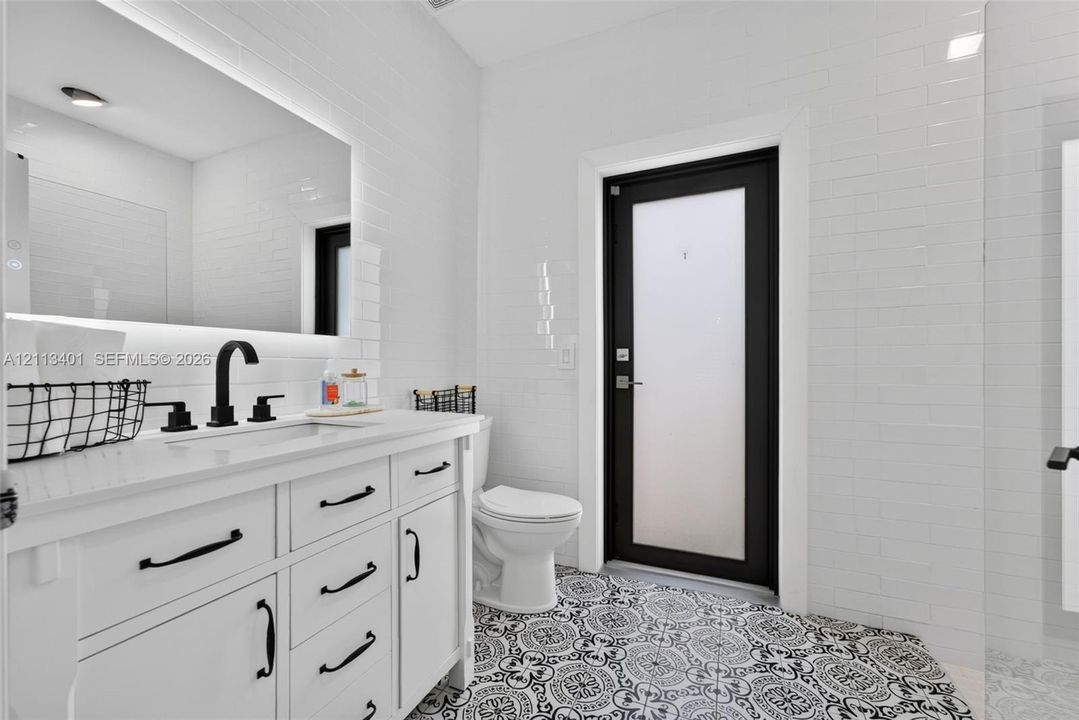 White vanity with a light-toned countertop, matte black hardware, integrated backsplash, backlit mirror, and patterned tile flooring