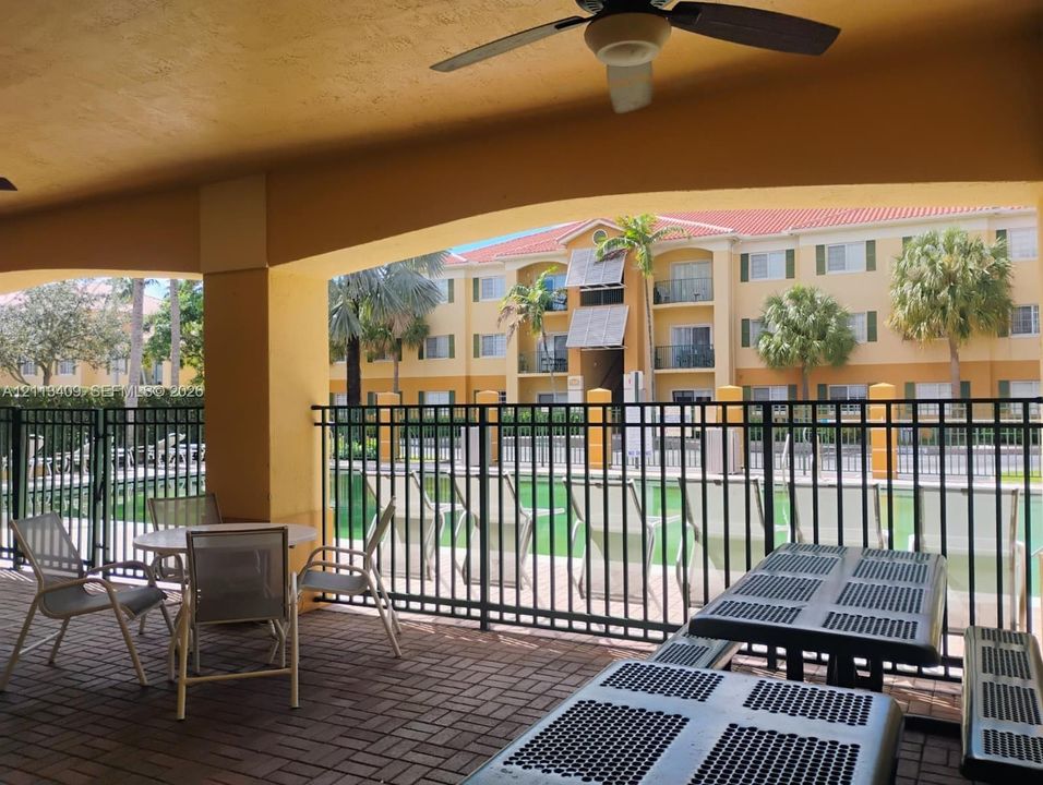 Covered patio featuring brick pavers, a ceiling fan, and a metal perimeter fence overlooking an outdoor swimming pool