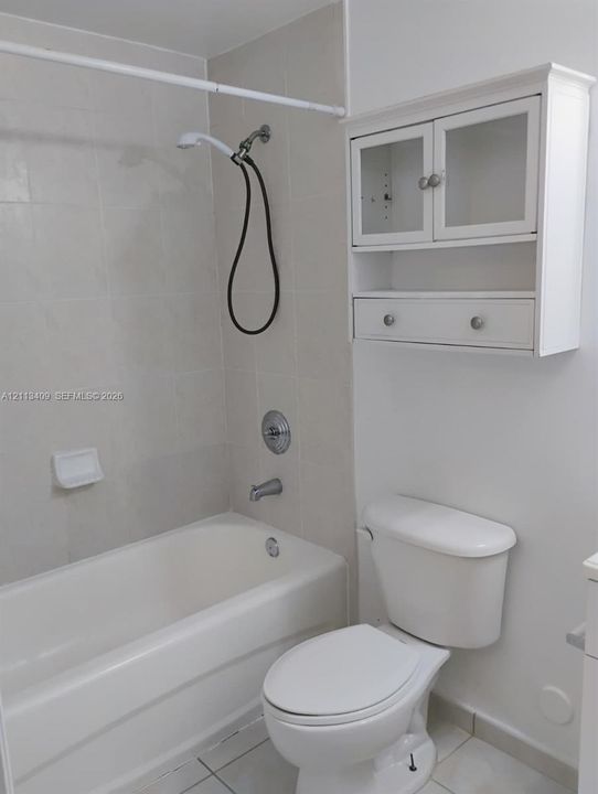 Bathroom featuring a tiled tub surround, handheld showerhead, white toilet, and white wall-mounted cabinet with glass-front doors