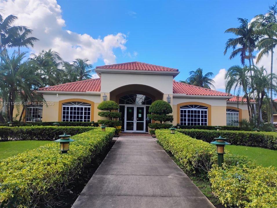 Stucco exterior featuring a terracotta tile roof, arched entryway with double-door entry, multi-pane windows, and a paved walkway flanked by manicured shrubbery and outdoor lighting fixtures