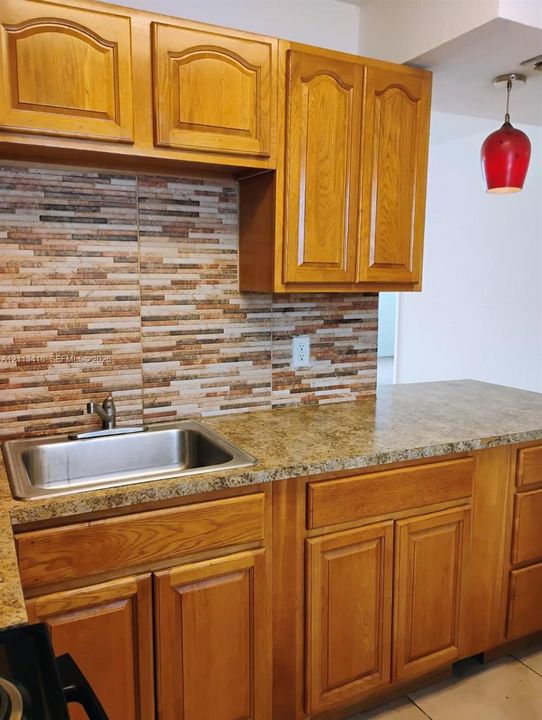 Kitchen featuring wood cabinetry, stone-finish countertops, stacked-stone backsplash, stainless steel drop-in sink, and a pendant light fixture
