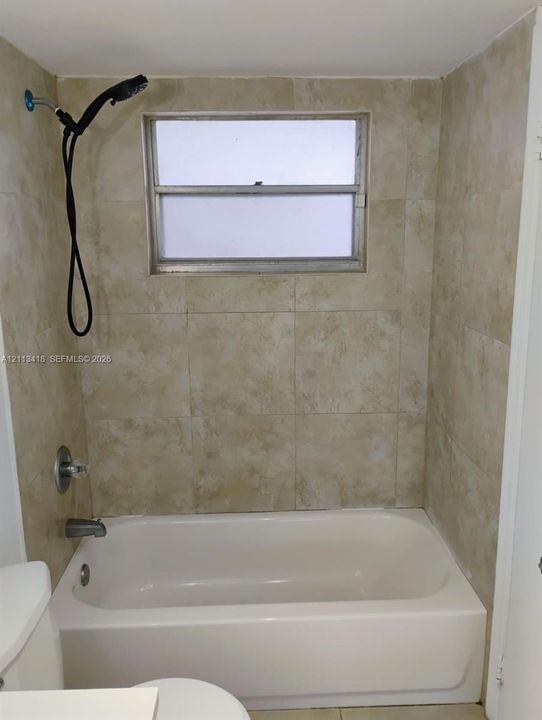 Bathtub enclosure featuring neutral-toned wall tile, a single-handle shower valve, a wall-mounted handheld showerhead, and an operable window