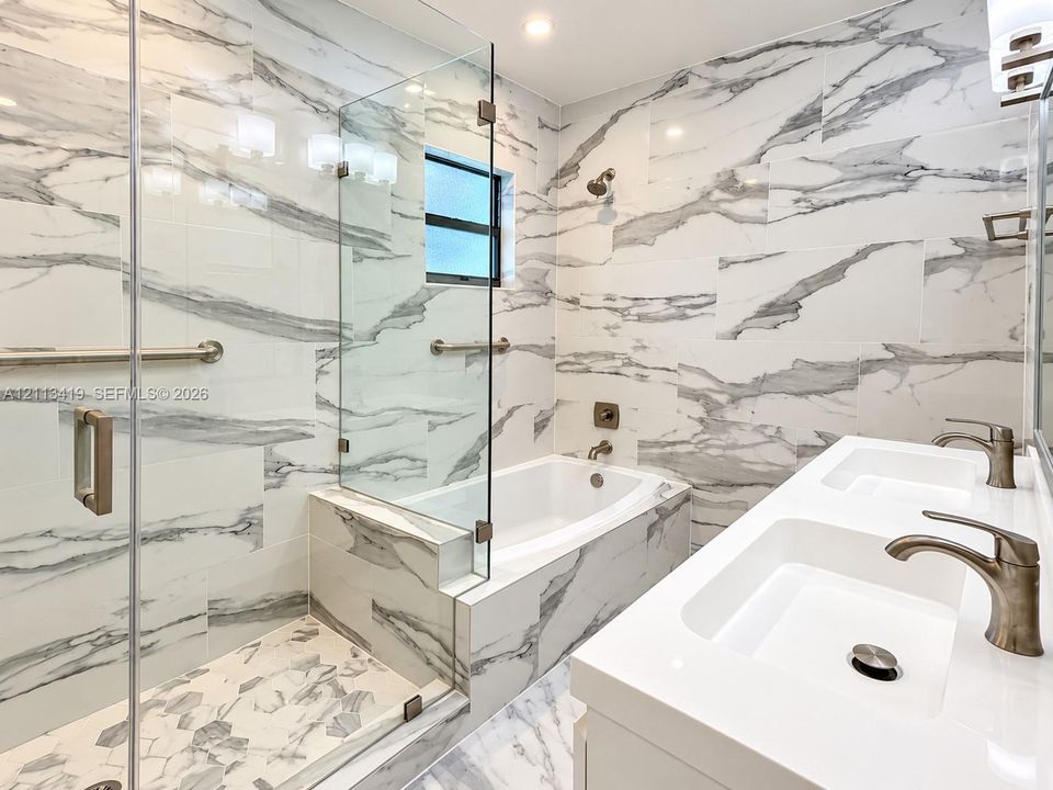 Full-height, large-format tile surround with stone-finish patterning, glass-enclosed walk-in shower, soaking tub, dual-basin vanity with polished metal fixtures, and recessed lighting