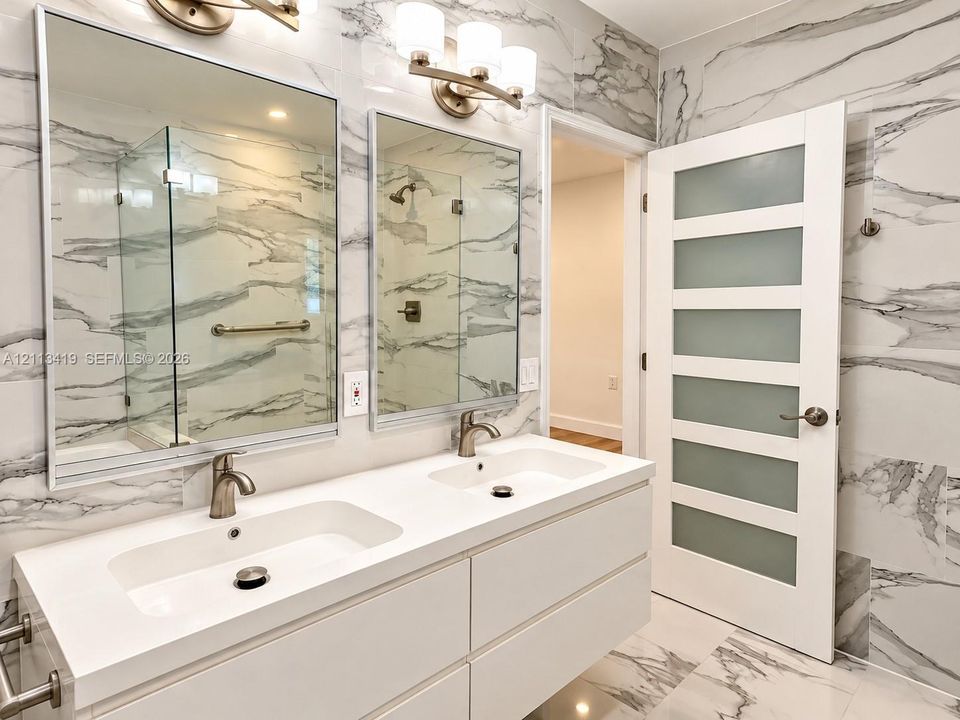 Dual-sink floating vanity with stone-composite countertop, full-height wall tiling in a marbled finish, rectangular mirrors, and a five-panel door with frosted glass inserts