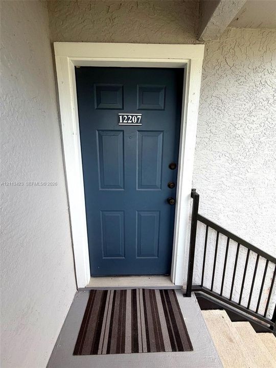 Exterior entryway featuring a solid blue door with six panels, white door casing, and a black metal handrail