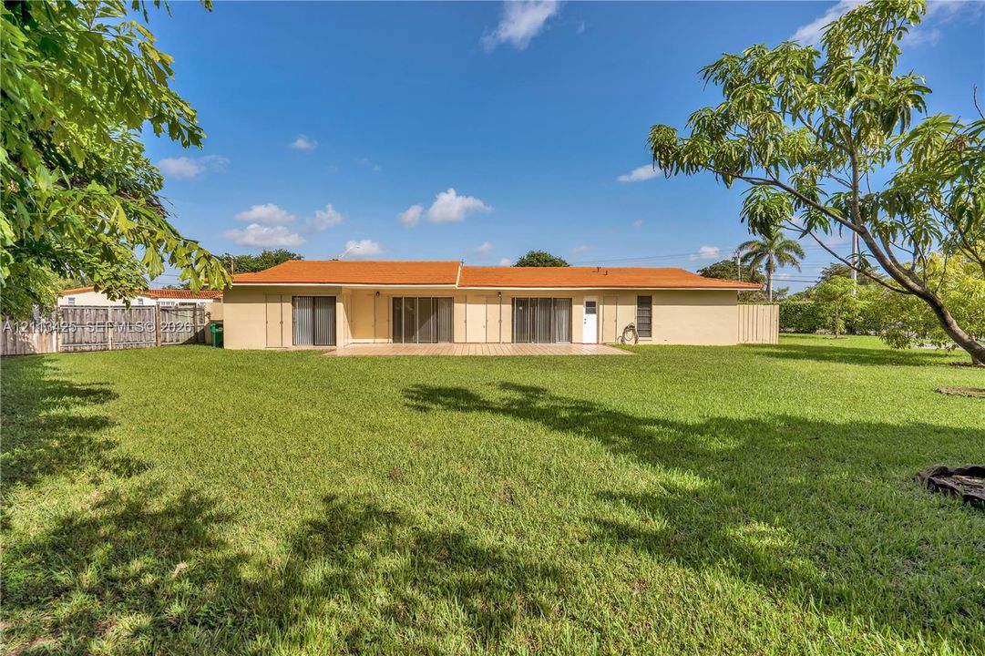 Spacious rear exterior featuring a manicured lawn, paved patio area, neutral-tone exterior finish, and mature trees
