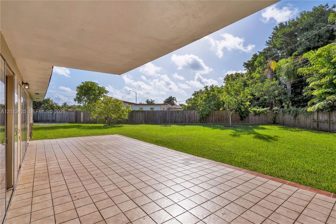Covered patio featuring ceramic tile flooring, expansive grass lawn, mature trees, and perimeter wood fencing