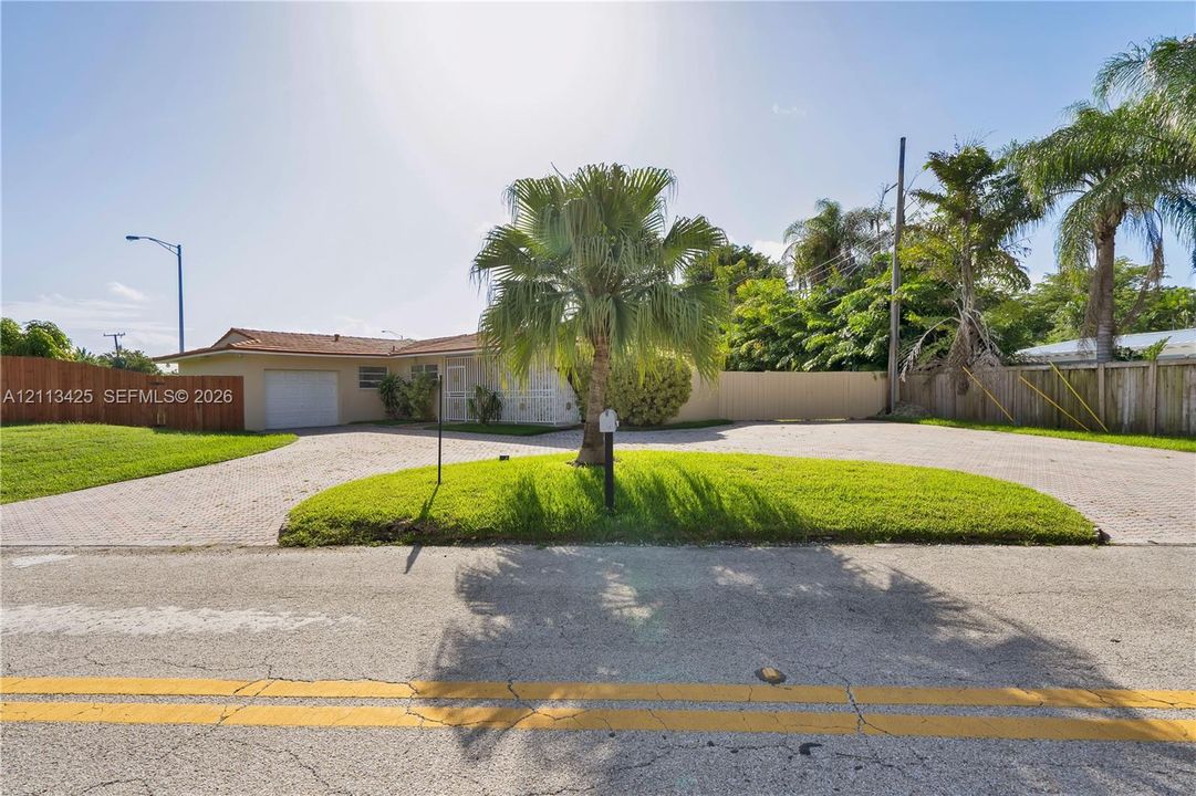 Exterior facade featuring a paved circular driveway, single-car garage, manicured lawn, palm tree, and perimeter wood fencing