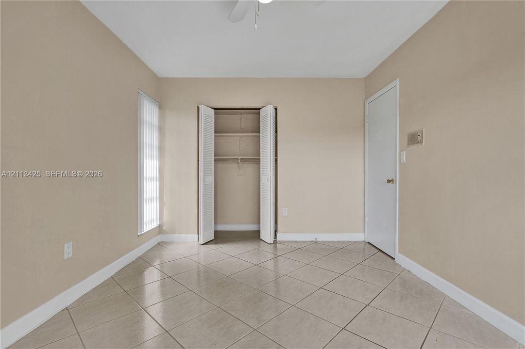 Neutral-toned interior room featuring diagonal tile flooring, a closet with louvered bi-fold doors and built-in shelving, vertical window blinds, and a ceiling-mounted fan
