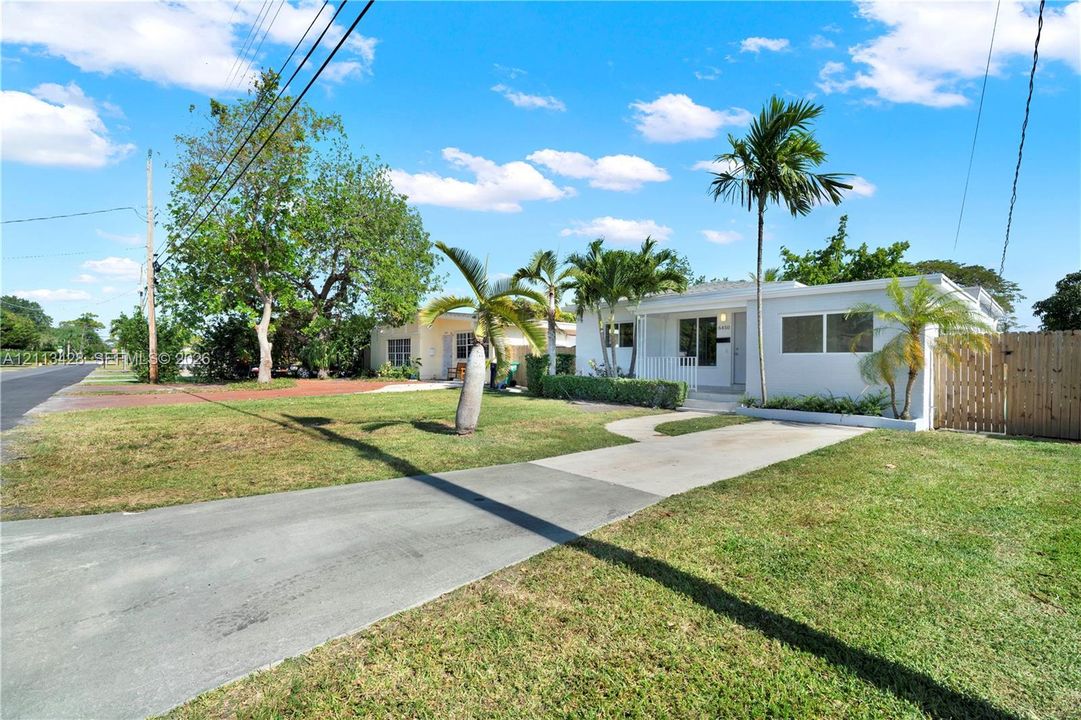 Single-story exterior featuring a light-toned stucco facade, concrete driveway, expansive grass lawn, mature palm trees, and a wooden privacy fence
