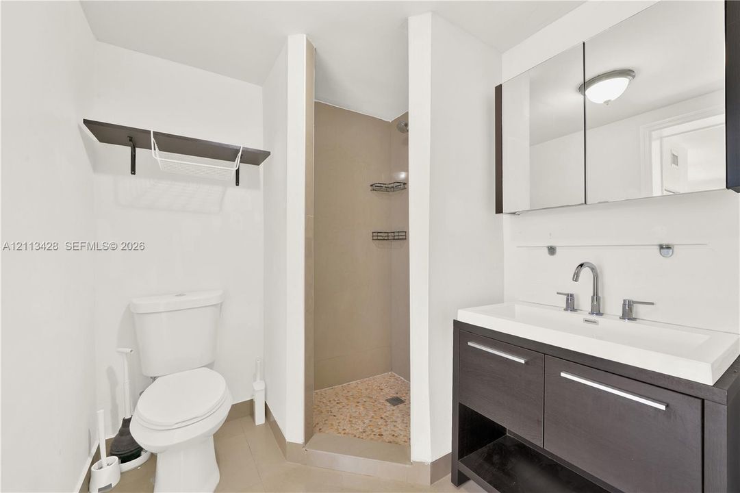 Bathroom featuring a vanity with dark cabinetry, a rectangular sink with a chrome faucet, a mirrored medicine cabinet, a walk-in shower with pebble-stone flooring, and a wall-mounted shelving unit