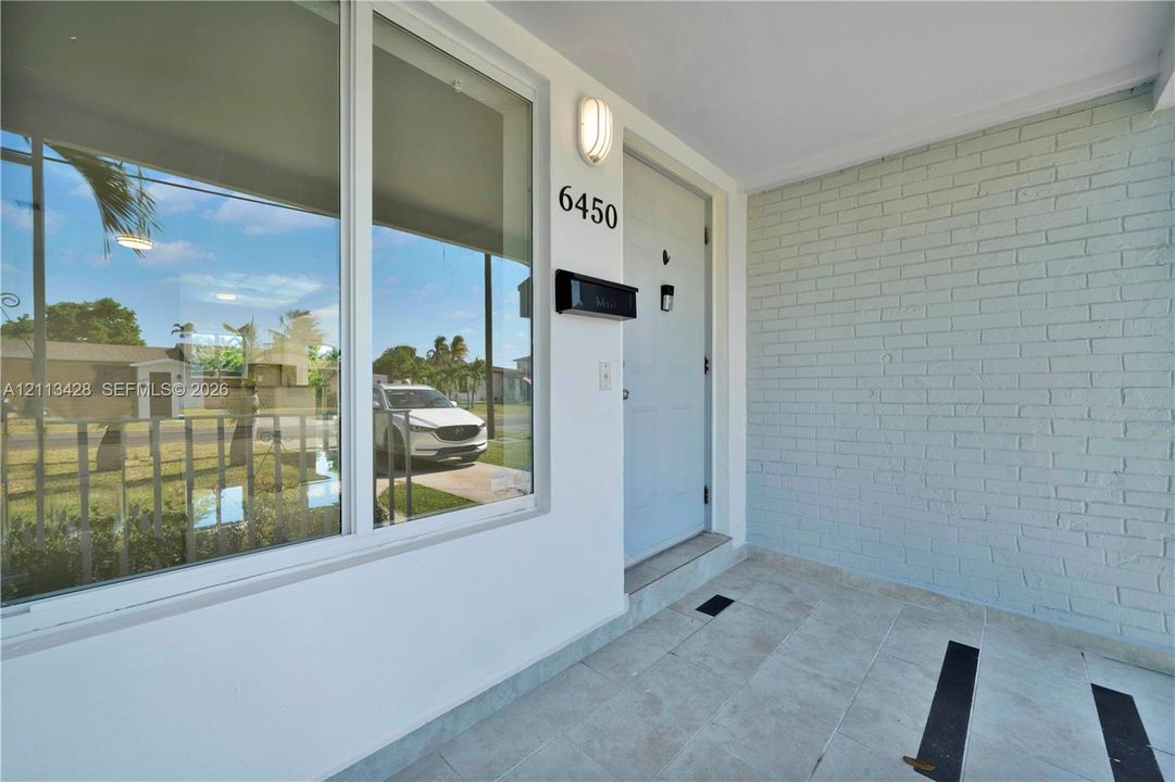 Covered entryway featuring a painted brick accent wall, large picture window, stone-finish tile flooring, and wall-mounted exterior lighting