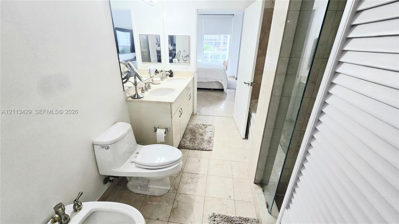 Bathroom featuring a dual-sink vanity with light-toned cabinetry, stone countertop, wall-to-wall mirror, glass-enclosed shower, and tile flooring