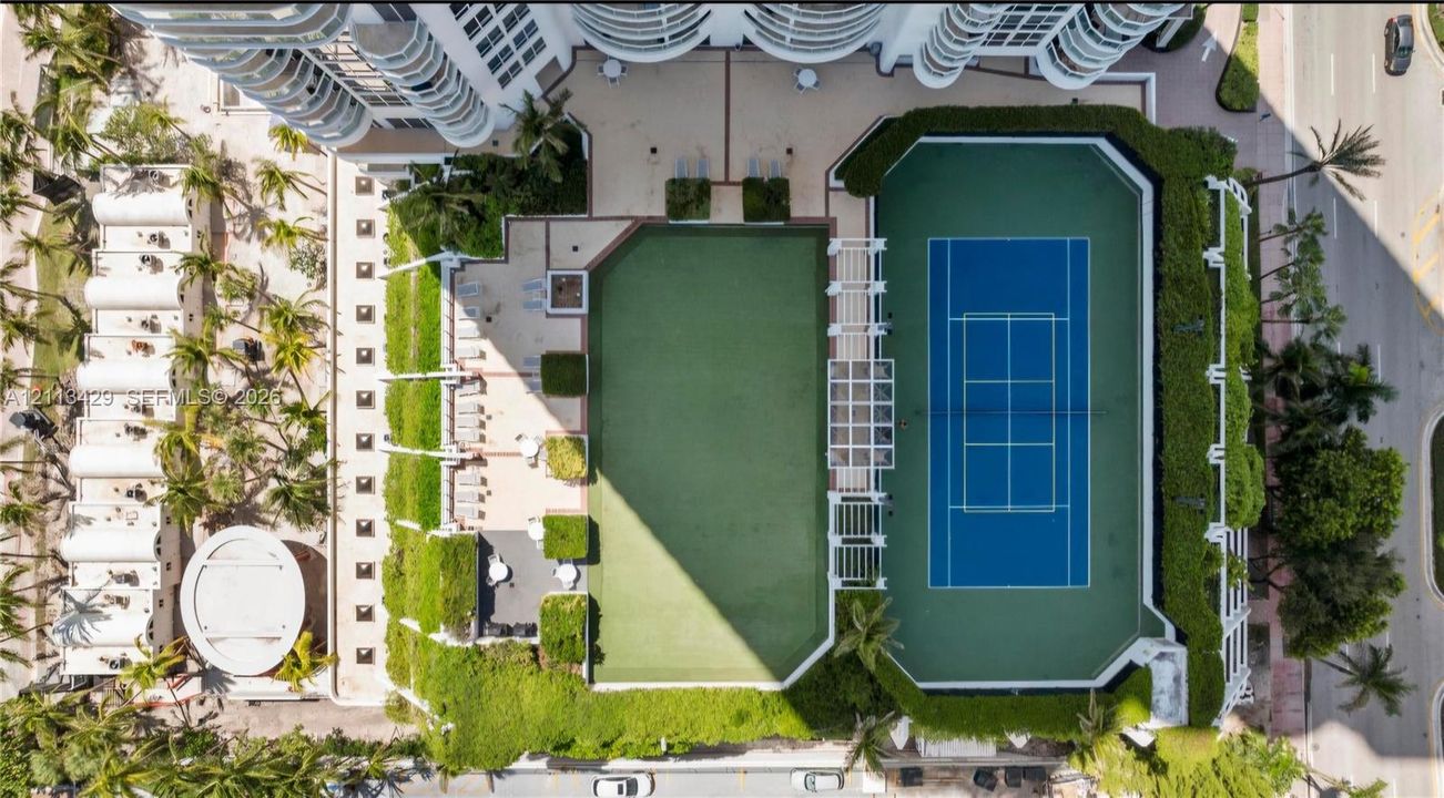 Aerial recreation deck featuring a hard-court tennis surface, synthetic turf lawn, white pergola structure, and landscaped perimeter hedging