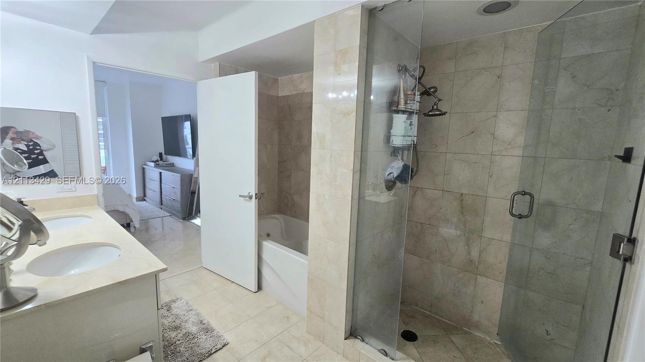 Dual-sink vanity with light-toned countertop, soaking tub, glass-enclosed shower with stone-tile surround, and tiled flooring