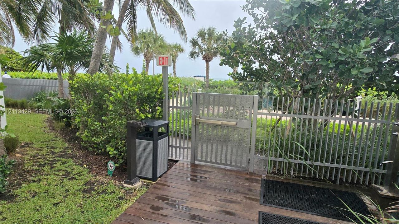 Outdoor area featuring a wood-finish deck, metal perimeter fencing with a gated entry, manicured landscaping, and mature palm trees