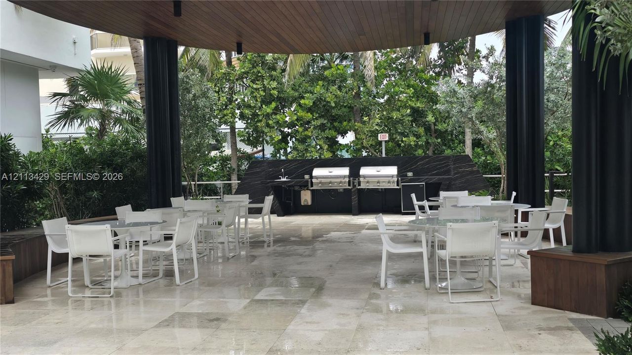 Covered outdoor kitchen featuring stone flooring, a dark stone backsplash, dual stainless steel grills, and a wood-paneled ceiling