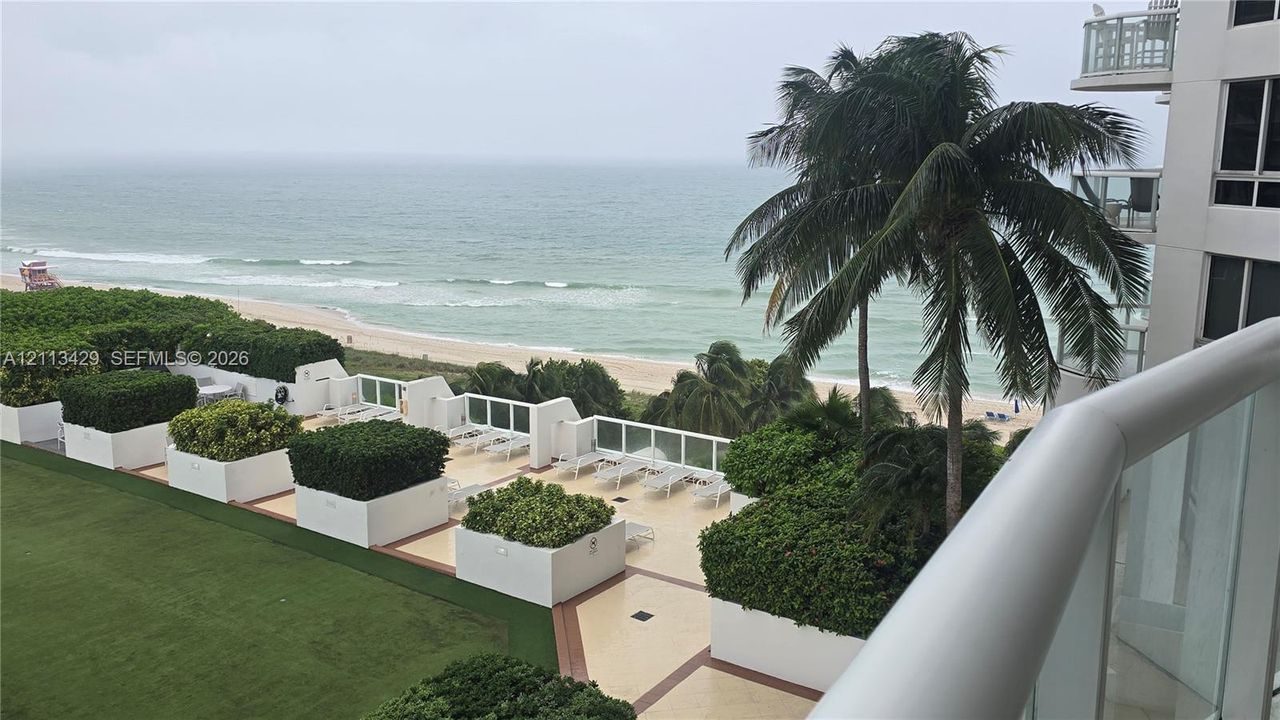Elevated terrace featuring white glass-panel railings, tile flooring, built-in planter boxes with mature greenery, and direct oceanfront scenery