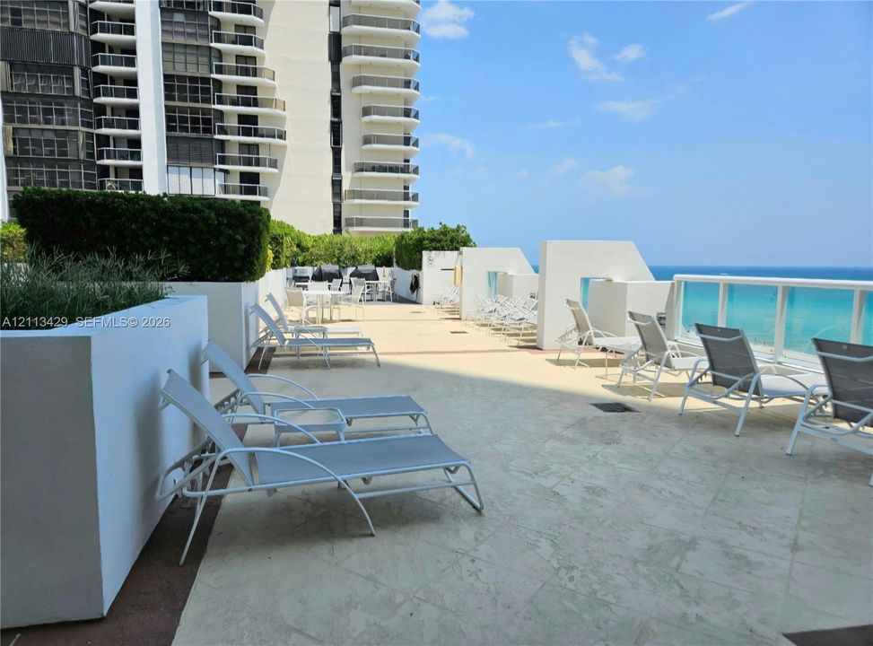 Expansive outdoor terrace featuring stone-tiled flooring, white masonry planters with manicured greenery, and glass-panel railing