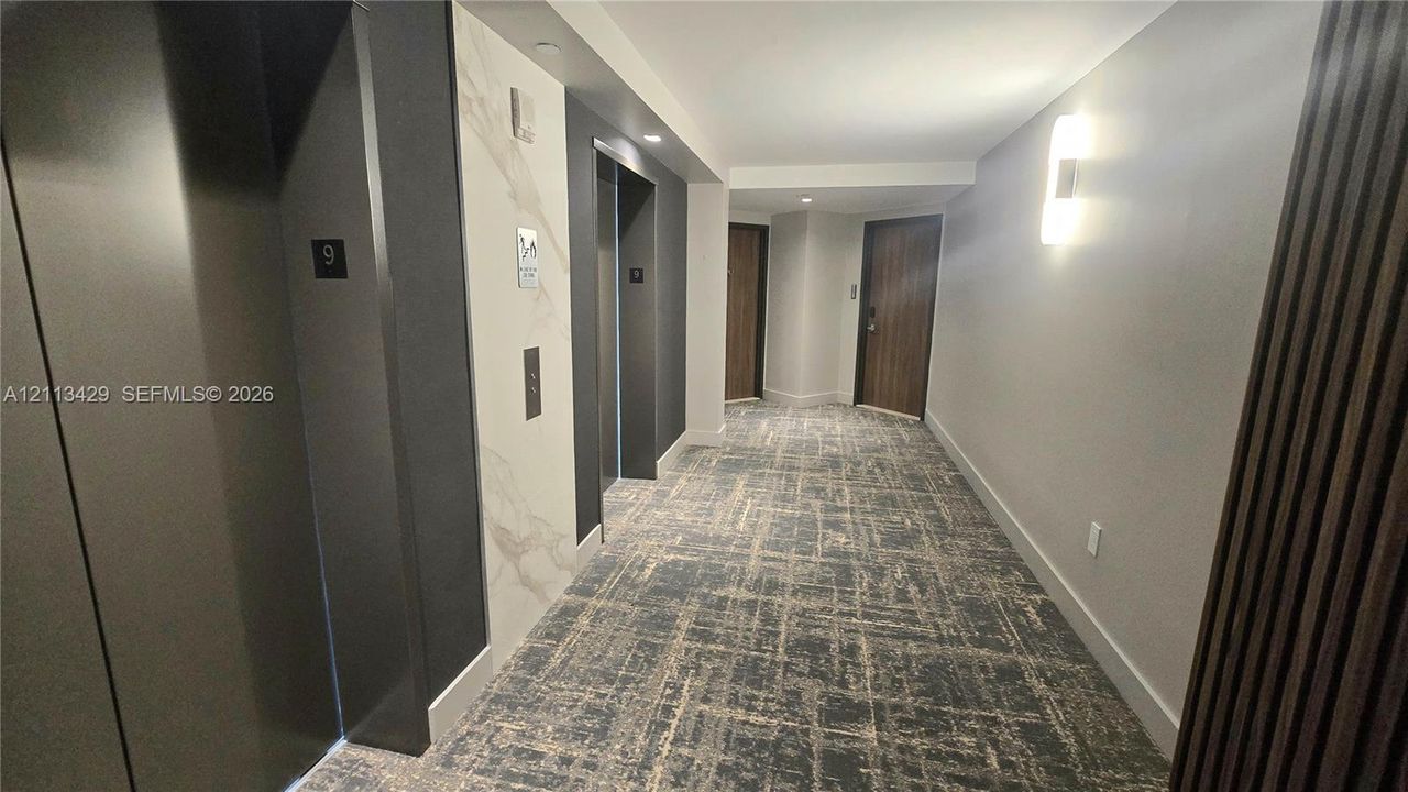 Hallway featuring patterned wall-to-wall carpeting, stone-look elevator surround, recessed lighting, and wall-mounted sconces