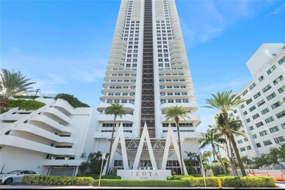 High-rise residential tower featuring a white facade, structural balconies, geometric entryway architecture, and established landscaping