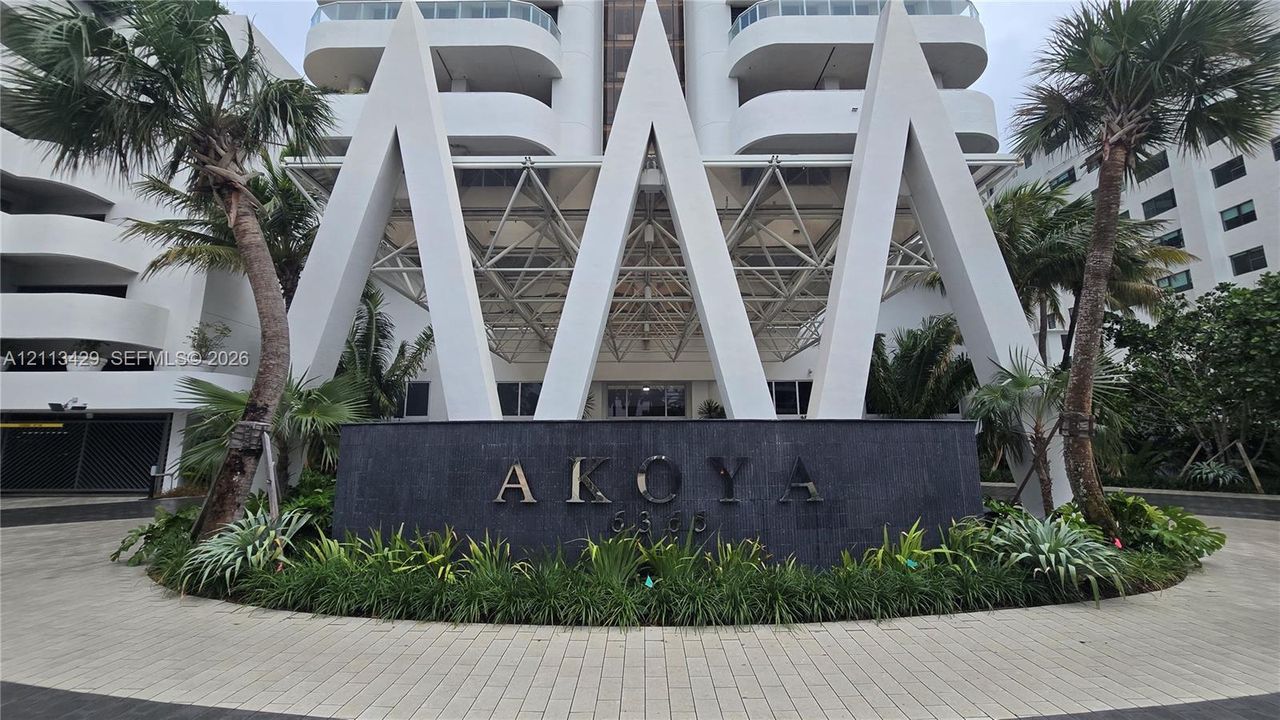 High-rise exterior featuring prominent white geometric structural detailing, a dark stone entrance monument with architectural signage, a paved circular motor court, and mature palm landscaping