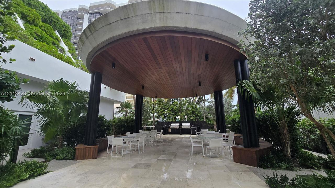 Circular outdoor pavilion featuring a wood-plank ceiling, black cylindrical columns, stone-tile flooring, and a built-in outdoor kitchen station