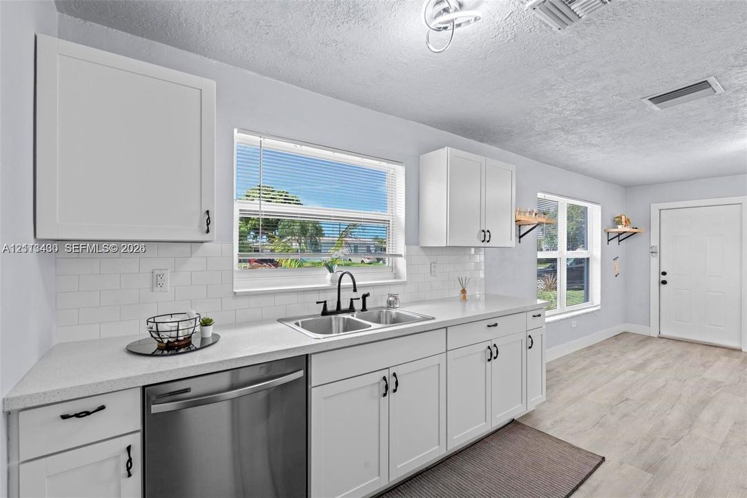 Kitchen featuring white shaker-style cabinetry, white subway tile backsplash, solid-surface countertops, dual-basin stainless steel sink with matte black hardware, and wood-finish flooring