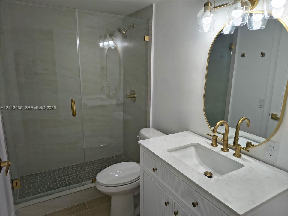 Bathroom featuring a white vanity with a light-toned stone countertop, brass-finish fixtures, a frameless glass-enclosed shower with a stone-tile surround, and an arched mirror with decorative overhead lighting