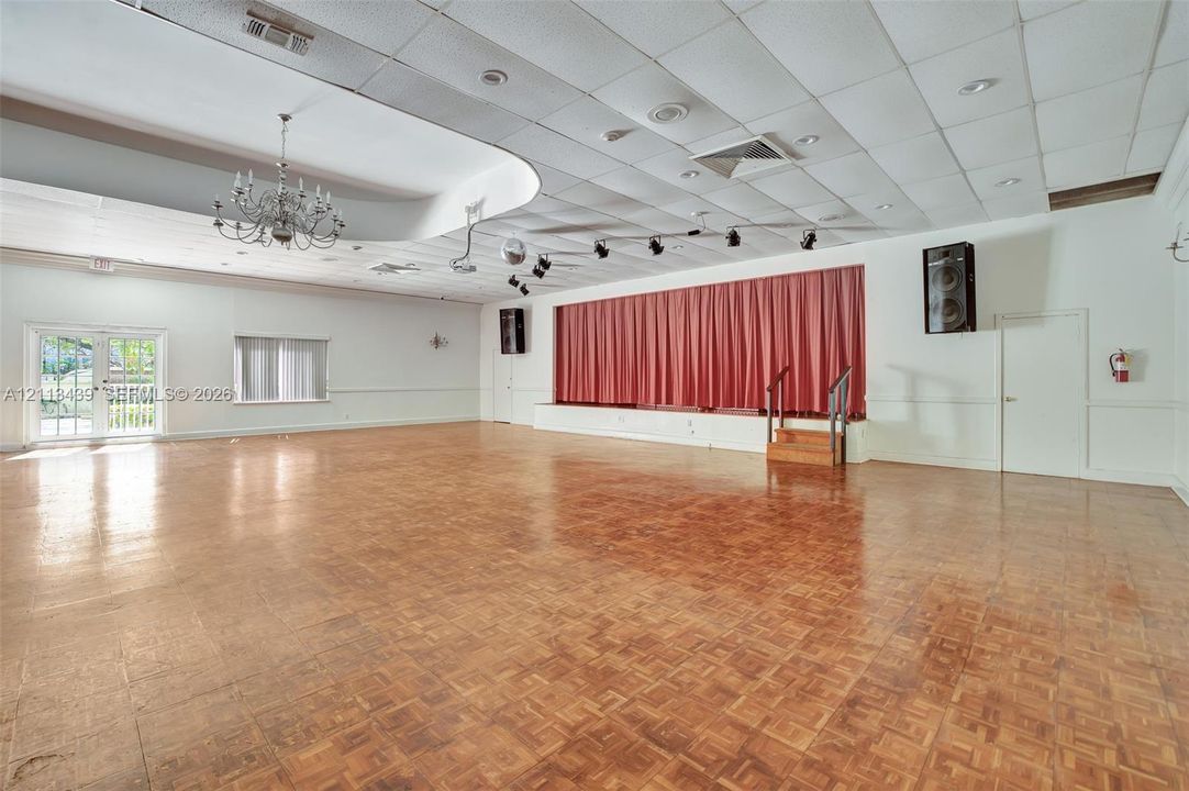 Spacious interior featuring wood-finish parquet flooring, a raised stage with drapery, an ornate chandelier, a suspended drop ceiling, and French doors