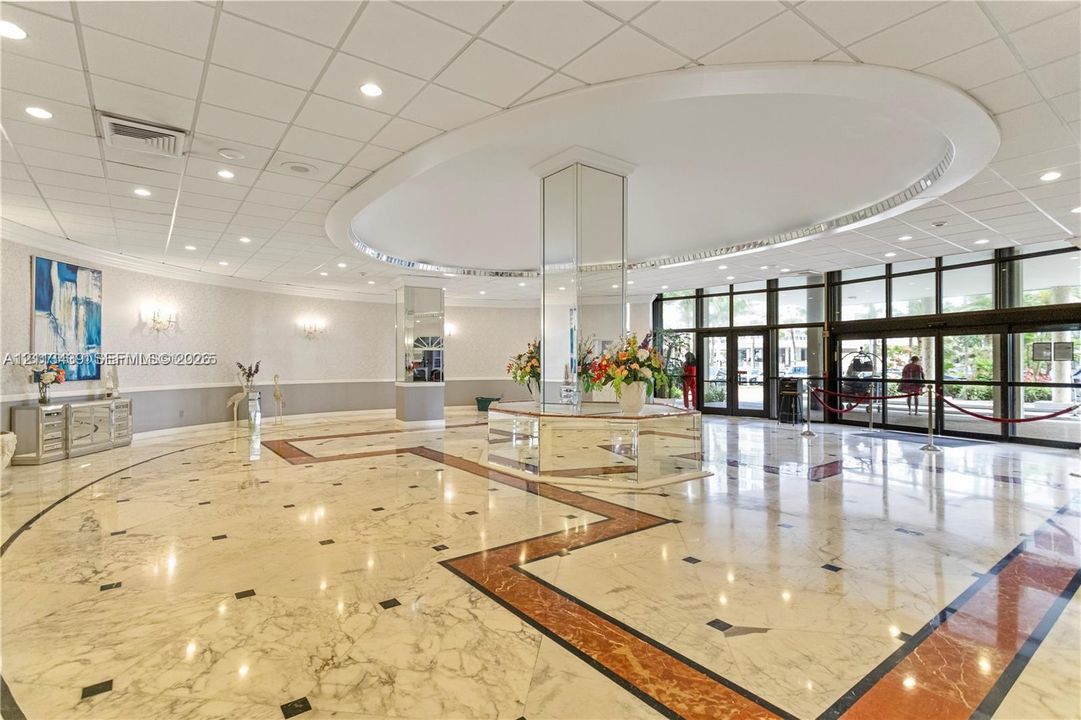 Expansive lobby featuring polished stone flooring with geometric tile inlay, a central mirrored support pillar, recessed lighting, drop ceiling design, and floor-to-ceiling glass entrance doors