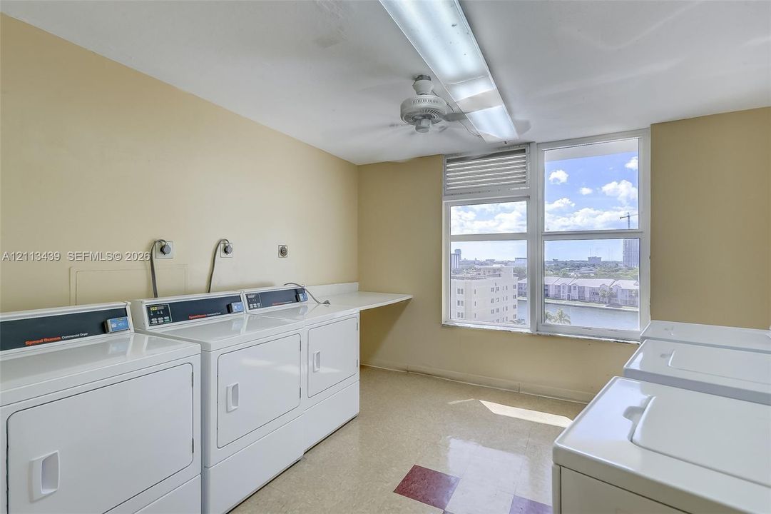 Utility room featuring multiple high-capacity washer and dryer units, expansive built-in workspace counter, large window with natural light, and ceiling-mounted lighting