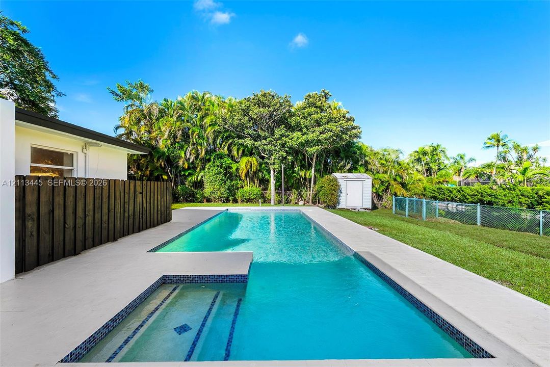 Swimming pool with an integrated sun shelf, mosaic tile waterline, concrete pool deck, dark wood-finish privacy fencing, and a detached storage shed